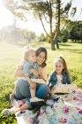 Joyeux jeune mère avec des petites filles mignonnes dans des vêtements similaires profiter ensoleillé journée d'été ensemble tout en étant assis sur la couverture sur pelouse herbeuse dans le parc — Photo de stock