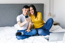 Deleitada madre étnica y adolescente con síndrome de Down sentado en la cama y tomando autorretrato en el teléfono inteligente - foto de stock