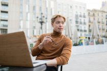 Freelancer masculino asiático Pensivo sentado com laptop à mesa no café de rua e trabalhando remotamente na inicialização enquanto olha para longe — Fotografia de Stock