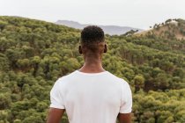 Задумчивый афроамериканец в модной одежде, стоящий на природе и любующийся видом горного леса — стоковое фото