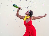 Entzückte Afroamerikanerin in Blumenkette trägt hellen Overall stehend mit Flasche Champagner vor weißem Hintergrund — Stockfoto