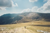 Отдаленный взгляд на неузнаваемого туриста, идущего по песчаной дороге в высокогорной долине во время похода в солнечный день в Уэльсе — стоковое фото
