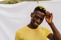 Bonito sorriso afro-americano masculino com flores amarelas no cabelo olhando para o fundo branco — Fotografia de Stock