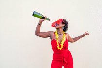 Cheerful African American female in red apparel and glasses holding bottle of alcoholic beverage during party on light background — Stock Photo