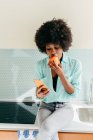 Modern beautiful African American female with smartphone in hand sitting on kitchen counter looking away at home and eating apple — Stock Photo