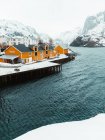 Casas amarelas e cais nevado localizados perto do mar ondulante contra montanhas no dia frio de inverno na aldeia costeira nas Ilhas Lofoten, Noruega — Fotografia de Stock