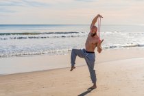 Vista lateral del atleta masculino sin camisa estirando los brazos con banda elástica mientras está de pie en una pierna haciendo ejercicio en la playa soleada vacía - foto de stock