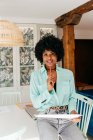 Современная успешная афроамериканка-фрилансер в стильном наряде с афро-волосами улыбается, глядя в камеру, сидя за столом и читая документ дома — стоковое фото
