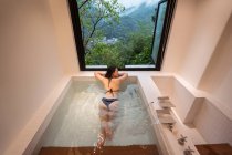 Señora étnica joven y relajada en traje de baño que se encuentra en el baño onsen japonés en el balneario junto a la ventana con vista a las montañas y árboles verdes - foto de stock