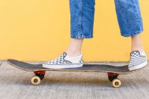 Cropped unrecognizable female skater with skateboard standing on walkway with colorful yellow wall on the background in daytime — Stock Photo