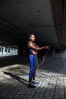 Vista laterale della bella donna afroamericana in abbigliamento sportivo che tiene la corda di salto e guarda la fotocamera mentre si trova sul marciapiede sulla strada della città — Foto stock