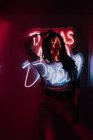 Junge hispanische Frau berührt Haare und blickt in die Kamera, während sie sich in der Nähe von Neon an die Wand lehnt — Stockfoto