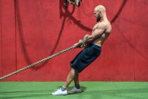 Vista lateral do desportista forte puxando corda com pesos pesados durante o treino intenso no ginásio contemporâneo — Fotografia de Stock