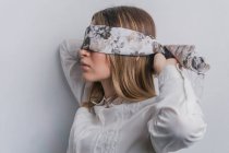 Side view of young fair haired female in elegant blouse covering eyes with folded silk scarf with floral ornament against white background — Stock Photo
