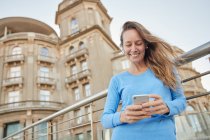 From below of smiling adult lady in casual clothes standing near fence and old building while surfing on phone in city district in daylight — Stock Photo