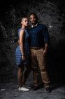 Zarter ethnischer Mann umarmt Frau auf dunklem Hintergrund im Studio und blickt in die Kamera — Stockfoto