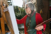 Seitenansicht von gealterten Künstlerinnen in Schutzmaske Malerei auf Leinwand im Hinterhof an sonnigen Tag im Sommer — Stockfoto