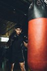 Joven centrado hombre asiático entrenamiento boxeo realizar golpes mientras se ejercita con saco de boxeo pesado en un gimnasio moderno - foto de stock