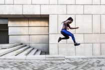 Vista lateral de la musculosa mujer afroamericana en ropa deportiva saltando alto en el aire mientras hace ejercicio cerca de la pared del edificio moderno en la calle de la ciudad - foto de stock