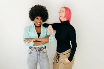 Cheerful young pink haired woman and African American girlfriend in stylish outfit while having fun together on white background — Stock Photo