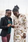 Традиционные улыбчивые афроамериканские дамы с прической проводят время вместе за мобильным телефоном в парке в яркий день — стоковое фото