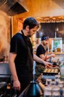 Бічний вид на шеф-кухаря в чорній формі і бандана куховарство азіатська страва називається ramen в сучасному кафе. — стокове фото
