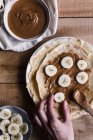 Top view of anonymous person putting pieces of fresh banana on palatable crepes with nut butter on timber table — Stock Photo