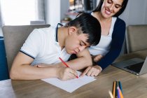 Етнічний хлопчик-підліток з синдромом Дауна малює олівцями на папері, сидячи за столом з фрілансером, який працює на ноутбуці вдома — стокове фото