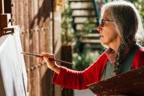 Side view of aged female artists in protective mask painting on canvas in backyard on sunny day in summer — Stock Photo