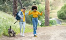 Full body young man holding hand and teaching cheerful black girlfriend to ride skateboard on countryside road near dog in nature — Stock Photo