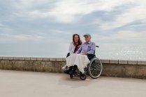 Зміст доросла дочка і батько літніх людей в інвалідному візку на набережній проти моря разом влітку в — стокове фото