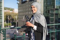 Femme entrepreneur musulmane joyeuse dans le hijab et avec café à emporter debout dans la rue — Photo de stock