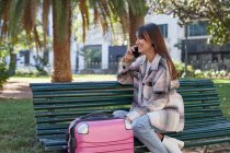 Positive young female traveler in stylish coat sitting on bench near suitcase and talking on mobile phone in urban park in spring day — Stock Photo