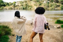 Back view of unrecognizable female friends with dog and skateboard standing near river and admiring nature during camping trip together in summer countryside — Stock Photo