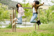 Couple of lesbian multiethnic females jumping above ground in woods and enjoying freedom together — Stock Photo