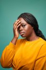 Unemotional grassoccio afroamericano femminile in maglione giallo e cappuccio toccare il viso e guardando la fotocamera mentre in piedi contro il muro blu — Foto stock