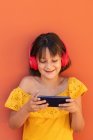 Зміст дитячого серфінгу в Інтернеті на мобільному телефоні, слухаючи пісню з бездротової гарнітури на помаранчевому фоні — стокове фото
