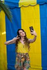 Mulher feliz com cabelo ondulado tirando selfie com telefone celular enquanto ri em dois fundo colorido na rua — Fotografia de Stock