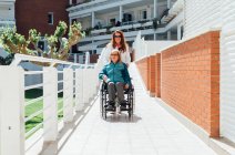 Fille adulte heureuse poussant fauteuil roulant avec mère âgée tout en profitant de flâner sur la journée ensoleillée — Photo de stock