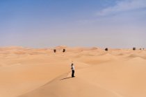Smiling young woman in casual clothes standing on sandy dune against desert during travel in Emirates and stretching arms — Stock Photo