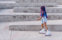 Side view of ethnic child in roller skates with colorful braids standing on staircase — Stock Photo