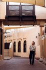 Full length joyful Asian female traveler looking away and smiling while walking along street against beige and brown facades of old buildings with arched windows in Doha — Stock Photo