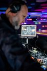 Nachdenklicher Mann mittleren Alters in Kopfhörern und schwarzer Freizeitkleidung, der sich konzentriert und professionellen DJ-Controller spielt, während er bei einem Konzert in einem zeitgenössischen Nachtclub gegen Laptop mit leerem Bildschirm auftritt — Stockfoto
