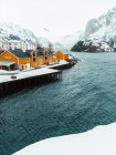 Cabañas amarillas y muelle nevado ubicado cerca del mar ondulante contra las montañas en el frío día de invierno en el pueblo costero en las Islas Lofoten, Noruega - foto de stock