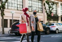Muslimische Freundinnen in Masken und mit Papiertüten überqueren die Straße, während sie nach dem Einkaufen in der Stadt spazieren gehen — Stockfoto