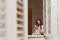 Femme joyeuse en pyjama debout près de la fenêtre et la navigation téléphone mobile à la maison — Photo de stock