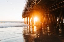 Сонячне світло, що світиться увечері, проникає в купи пристані Санта - Моніки з мирними океанічними хвилями, що йдуть на пляжі в Каліфорнії. — стокове фото