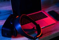 Angolo alto di scrivania in legno con netbook aperto vicino al telefono e occhiali VR in camera oscura con luci al neon — Foto stock