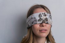 Young fair haired female in elegant blouse covering eyes with folded silk scarf with floral ornament against white background — Stock Photo