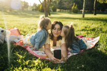 Mujer joven feliz y lindas hijas pequeñas tumbadas en la manta y tomando selfie en el teléfono inteligente mientras se divierten juntos en el prado verde en el parque de verano - foto de stock